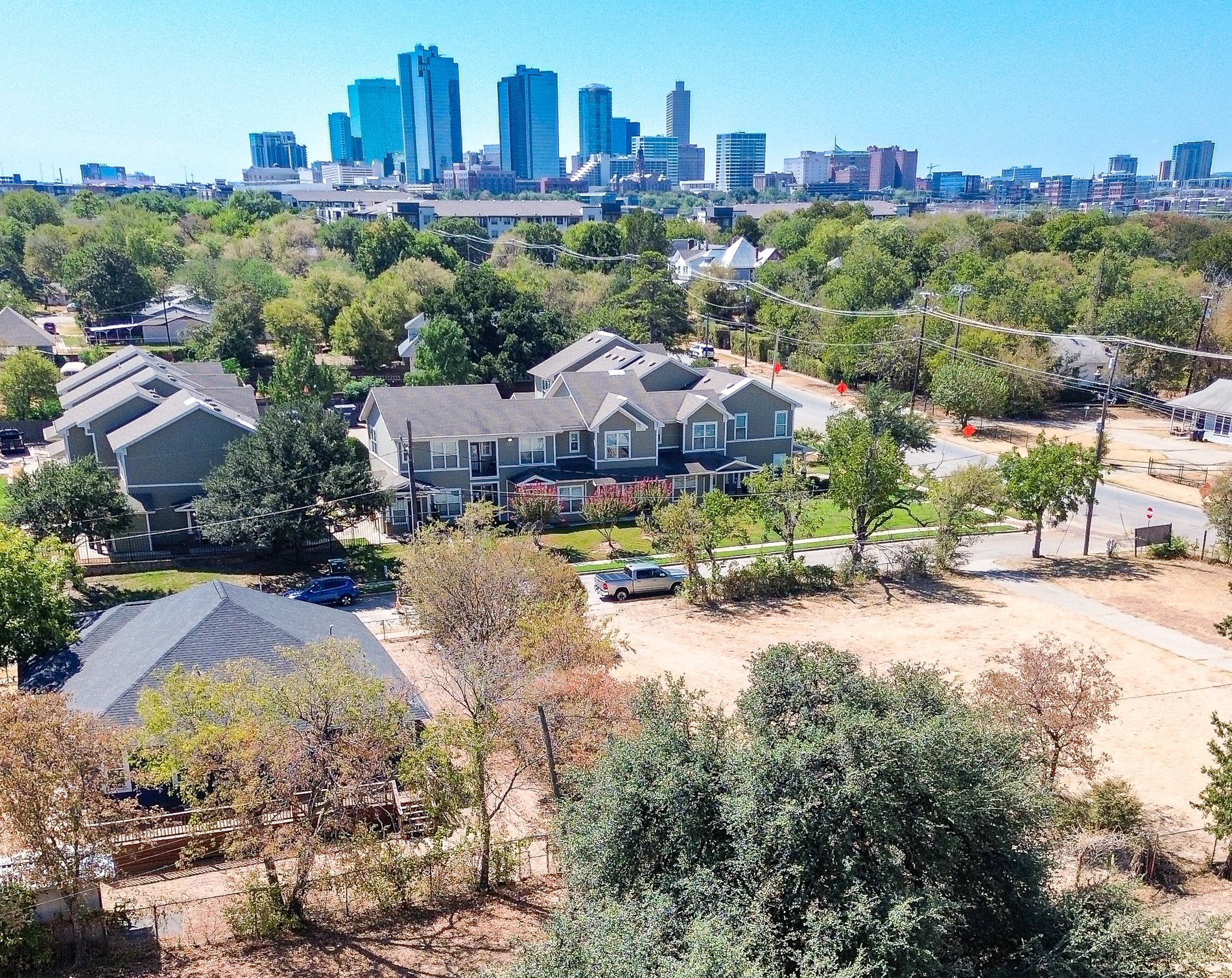 Aerial view looking southwest: the house with its back deck at lower left, the open ground and large oaks of Traders Oak Park filling the frame, townhomes and streets beyond, and the downtown Fort Worth skyline on the horizon.